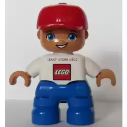 Duplo Figure Lego Ville, Child Boy, Blue Legs, White Top, Red Cap LEGO Logo and 'LEGO STORE UŠCE' on Front